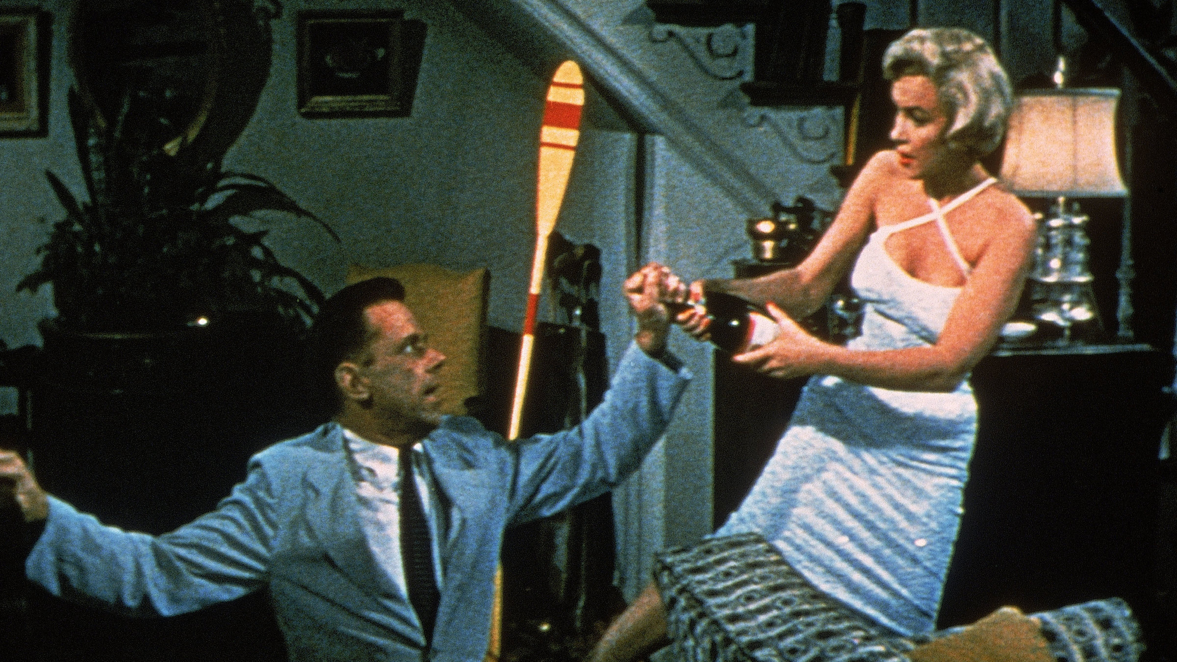 "Das verflixte 7. Jahr": Richard Sherman (Tom Ewell) trägt einen Anzug, kniet in einem Wohnzimmer auf dem Boden und blickt angestrengt nach oben, während seine linke Hand den Hals einer Sektflasche umfasst. Die Blondine (Marilyn Monroe), gekleidet in ein helles Cocktailkleid, schaut zu Sherman herunter, während sie die Sektflasche mit zwei Händen packt und versucht zu sich zu ziehen.