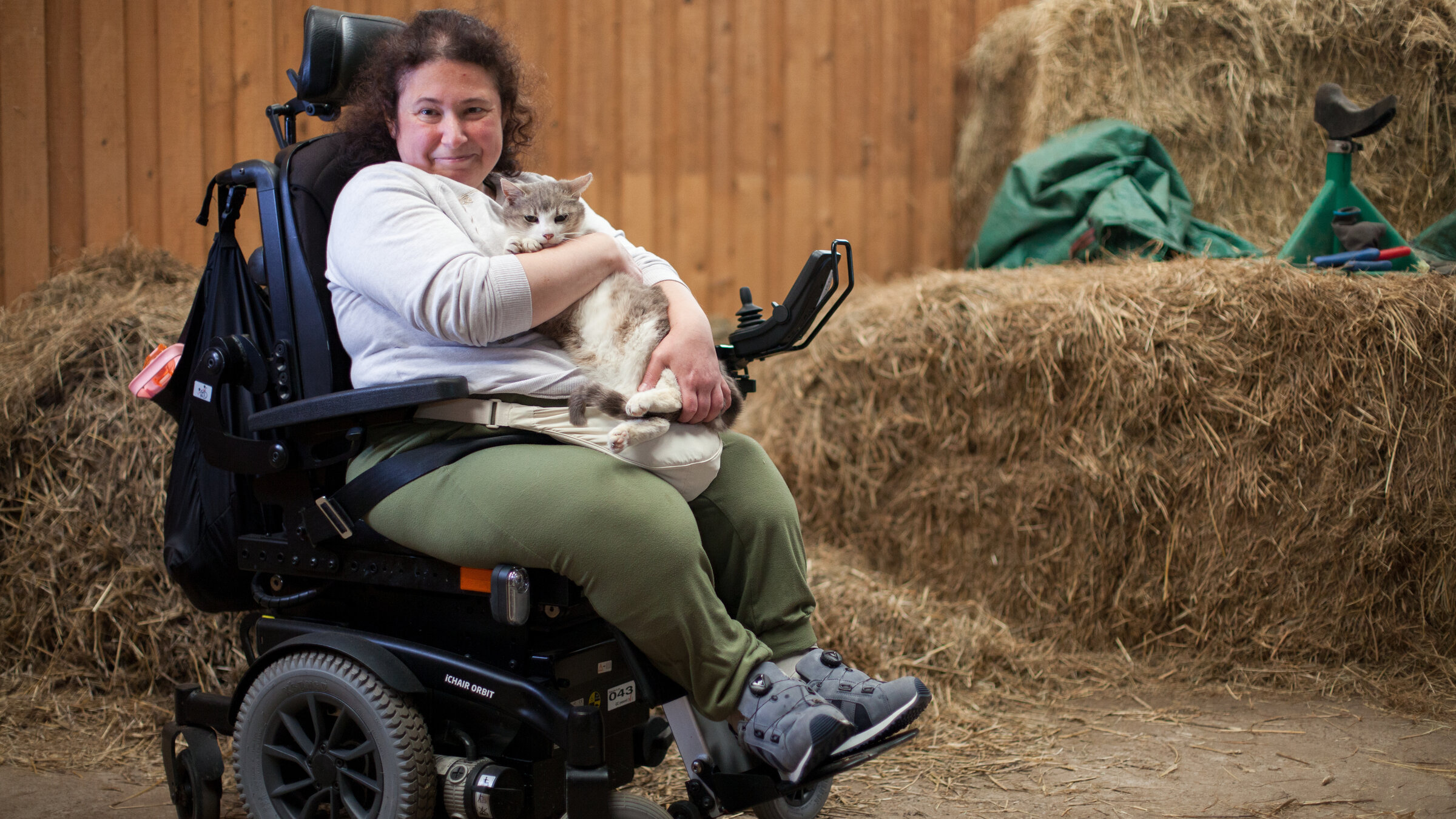 "Einfach Mensch - Barrierefrei unterwegs": Büsra Tutkunkardes sitzt im Rollstuhl im Stall des Reiterhofs Grüneberg in Brandenburg. Sie hält eine Katze im Schoß und schaut lächelnd in die Kamera.