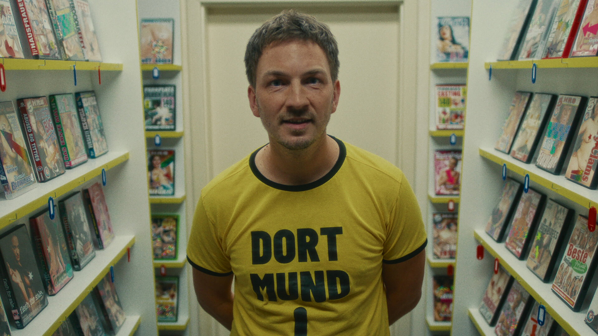 "Chabos - Warum liegt hier eigentlich Stroh?": Der erwachsene Peppi (Johannes Kienast) steht zwischen Regalen mit Porno-DVDs. Er trägt ein gelbes T-Shirt mit der schwarzen Aufschrift "Dort-Mund" und einem schwarzen Pfeil, der Richtung Schritt zeigt.