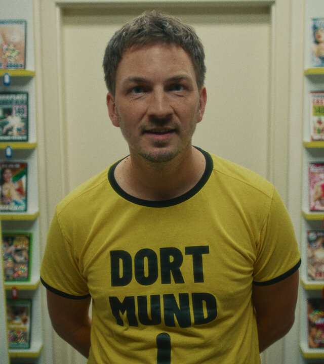 "Chabos - Warum liegt hier eigentlich Stroh?": Der erwachsene Peppi (Johannes Kienast) steht zwischen Regalen mit Porno-DVDs. Er trägt ein gelbes T-Shirt mit der schwarzen Aufschrift "Dort-Mund" und einem schwarzen Pfeil, der Richtung Schritt zeigt.