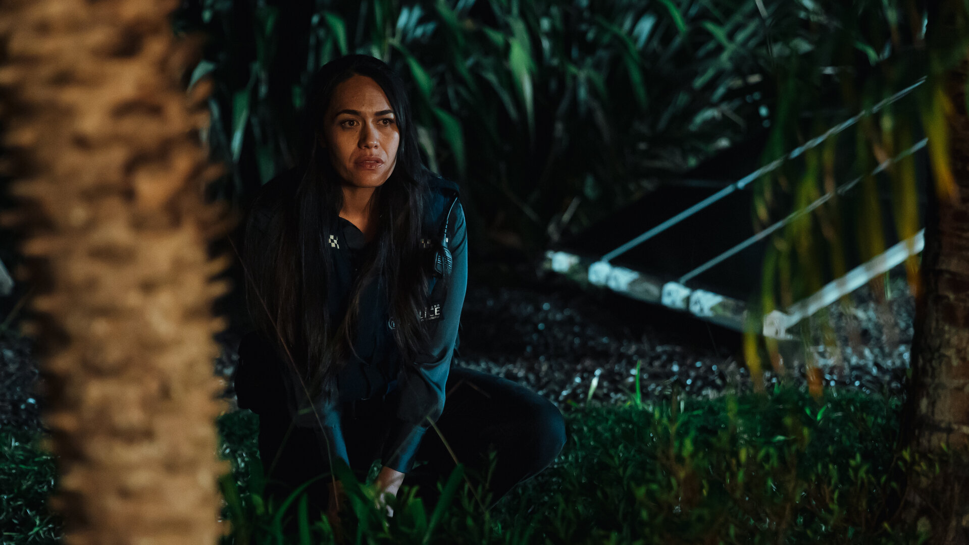 "Back Up - Auf Streife mit der Ex: Big Trouble in Little Waitote": DC Mihi Renata (Roimata Fox) sitzt im Dunklen neben einer Palme, im Hintergrund befinden sich grüne Büsche.
