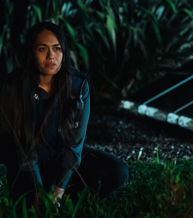 "Back Up - Auf Streife mit der Ex: Big Trouble in Little Waitote": DC Mihi Renata (Roimata Fox) sitzt im Dunklen neben einer Palme, im Hintergrund befinden sich grüne Büsche.