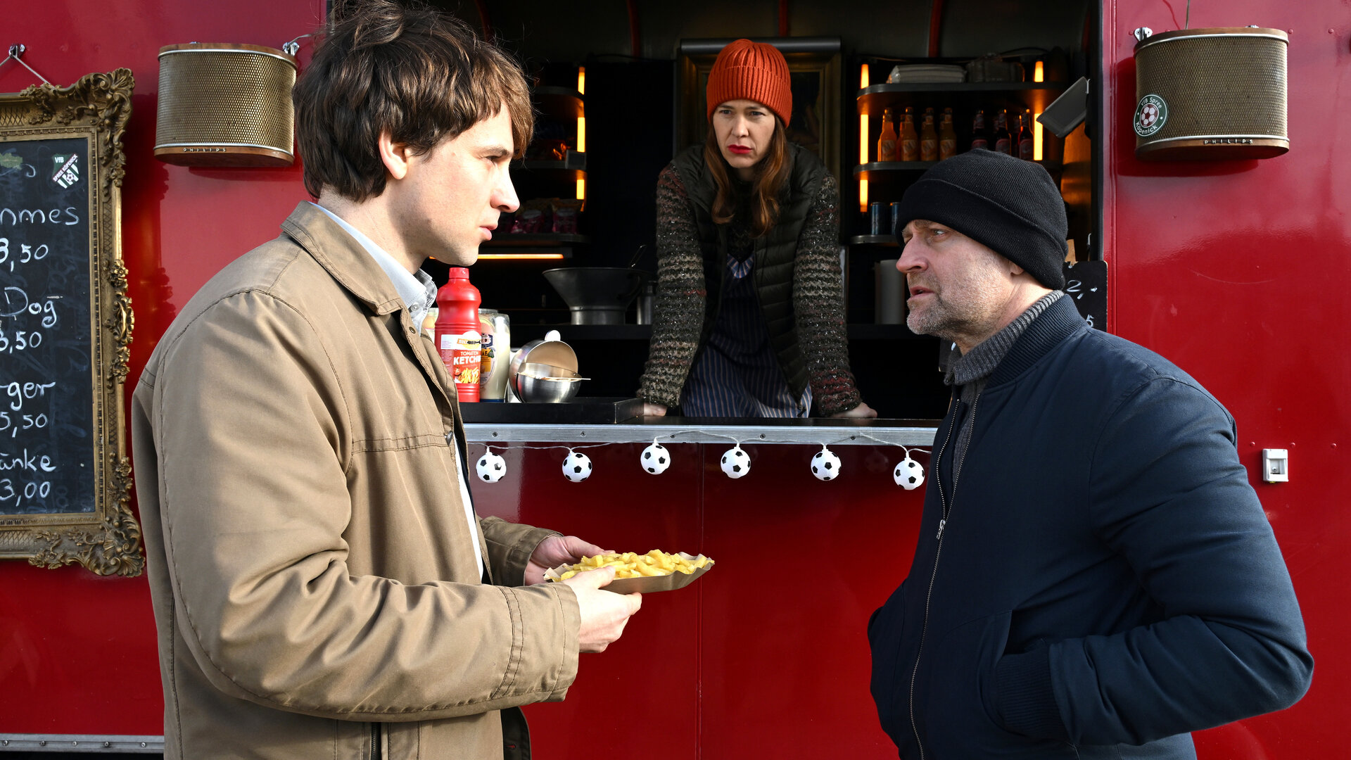"Jenseits der Spree - Hinter der Fassade": Benjamin Wortmann (Leonard Scheicher), Yvonne Moll (Meike Droste) und Robert Heffler (JÃ¼rgen Vogel) befinden sich an einem Imbissstand. Benjamin Wortmann hÃ¤lt eine Schale mit Pommes frites in den HÃ¤nden und steht Robert Heffler gegenÃ¼ber, der mit ihm im GesprÃ¤ch ist. Yvonne Moll lehnt sich aus der geÃ¶ffneten Verkaufsklappe des roten Imbisswagens und blickt zu den beiden.
