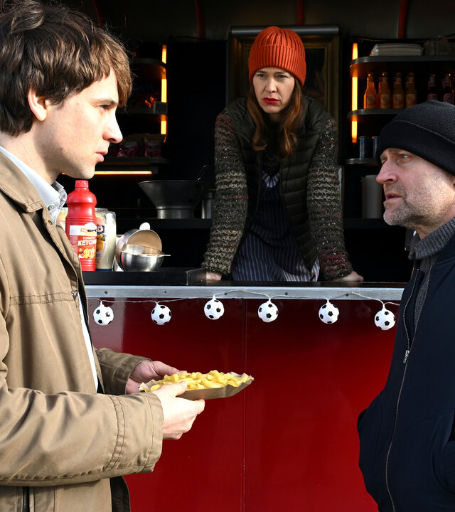 "Jenseits der Spree - Hinter der Fassade": Benjamin Wortmann (Leonard Scheicher), Yvonne Moll (Meike Droste) und Robert Heffler (JÃ¼rgen Vogel) befinden sich an einem Imbissstand. Benjamin Wortmann hÃ¤lt eine Schale mit Pommes frites in den HÃ¤nden und steht Robert Heffler gegenÃ¼ber, der mit ihm im GesprÃ¤ch ist. Yvonne Moll lehnt sich aus der geÃ¶ffneten Verkaufsklappe des roten Imbisswagens und blickt zu den beiden.