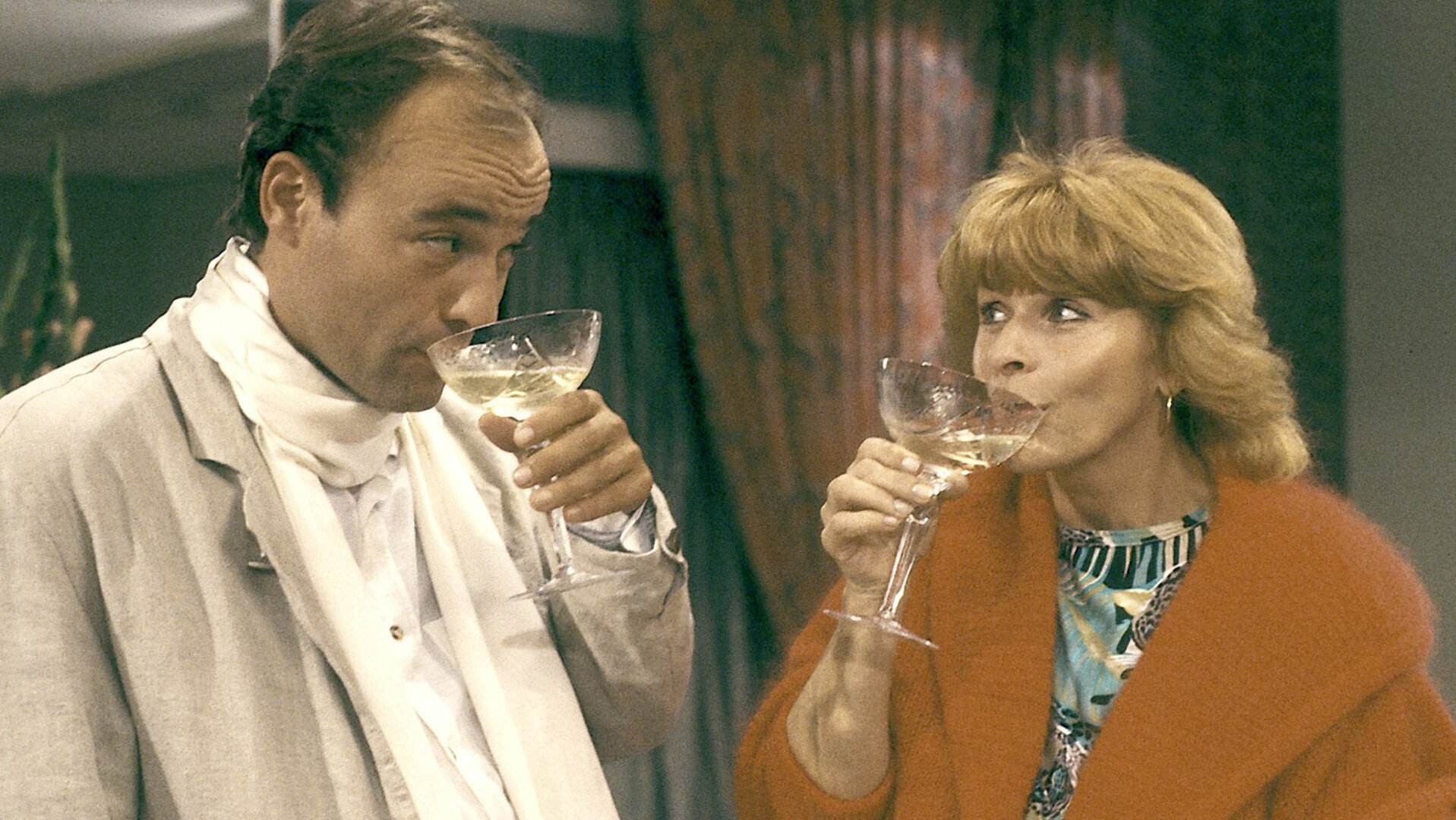 "Die schnelle Gerdi - Oktoberfest": Gerdi (Senta Berger) und Fernsehstar Sorbas (Heiner Lauterbach) stehen nebeneinander und trinken aus Schaumweingläsern.