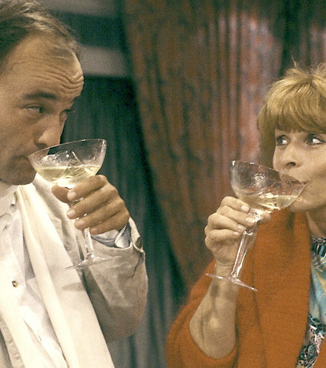 "Die schnelle Gerdi - Oktoberfest": Gerdi (Senta Berger) und Fernsehstar Sorbas (Heiner Lauterbach) stehen nebeneinander und trinken aus Schaumweingläsern.