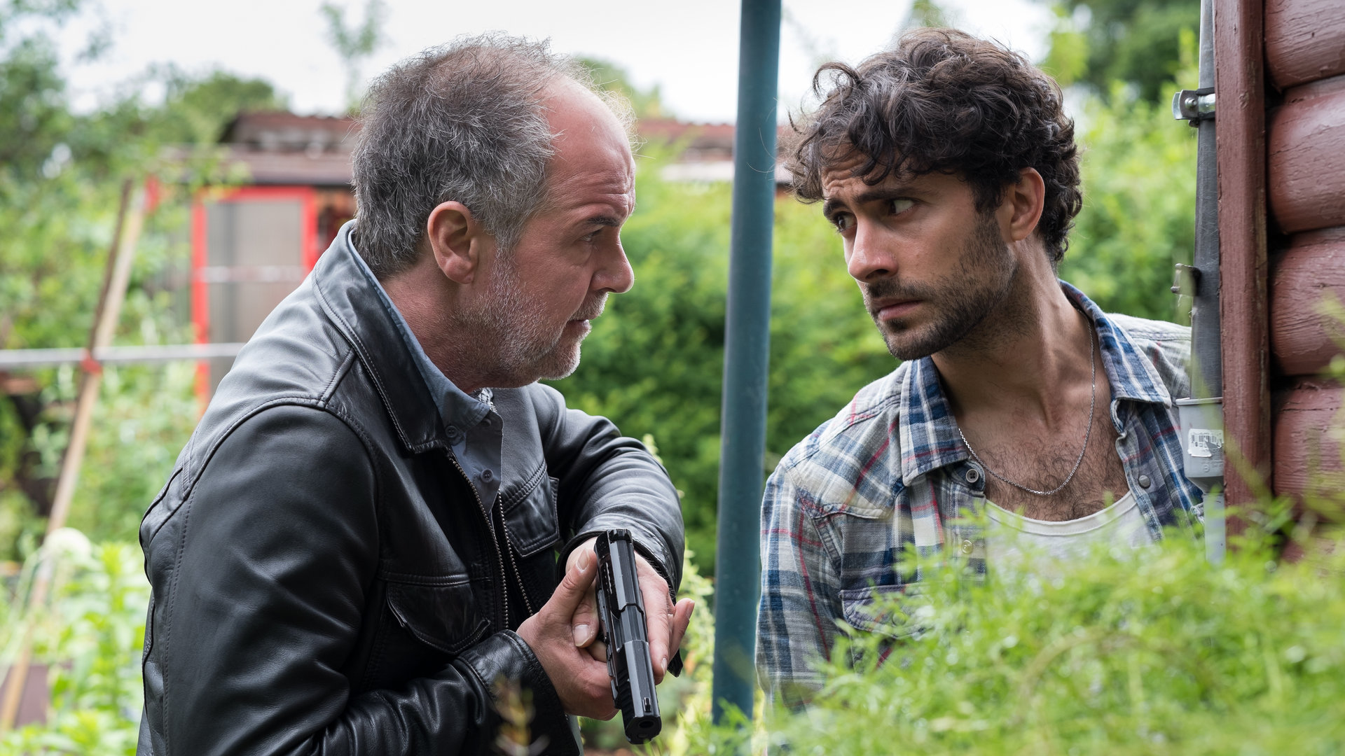 "SOKO Hamburg - Kleingarten Eden": Oskar Schütz (Marek Erhardt) und Max Nordmann (Garry Fischmann) stehen dicht nebeneinander hinter einem Gebüsch in einem Schrebergarten. Oskar Schütz blickt seinen Kollegen eindringlich an, dieser erwidert den Blick angespannt. Oskar hält seine gezückte Pistole mit beiden Händen vor die Brust, die Waffe zeigt leicht nach unten.