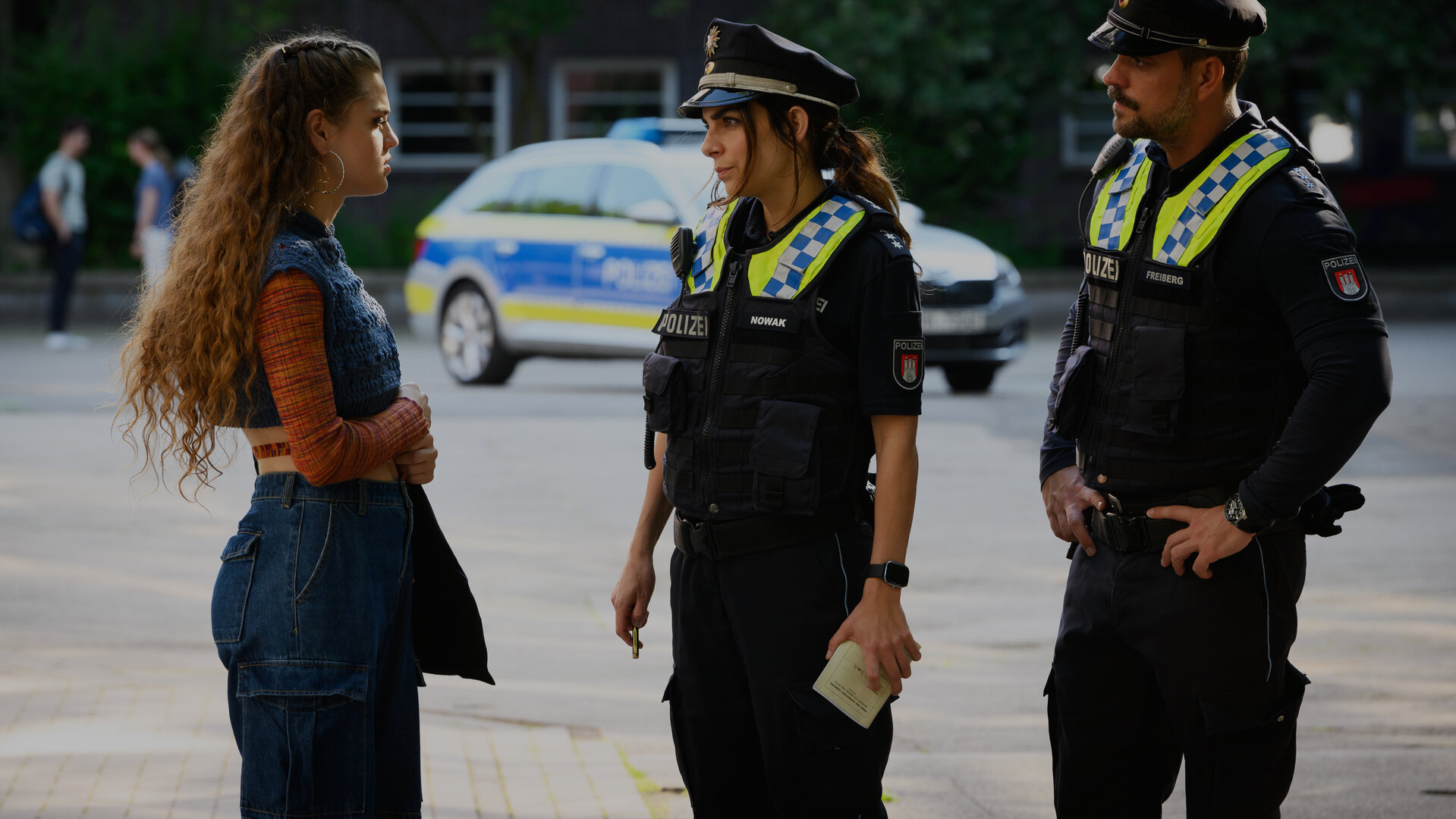 "Notruf Hafenkante - Wunschkind": Die Polizistin Isa Nowak (Lilli Hollunder) und ihr Kollege Kris Freiberg (Marc Barthel) stehen Lilly Grabow (Lilly Wiedemann) auf einem Schulhof gegenüber. Beide sehen sie an. In Hintergrund steht ein Streifenwagen.
