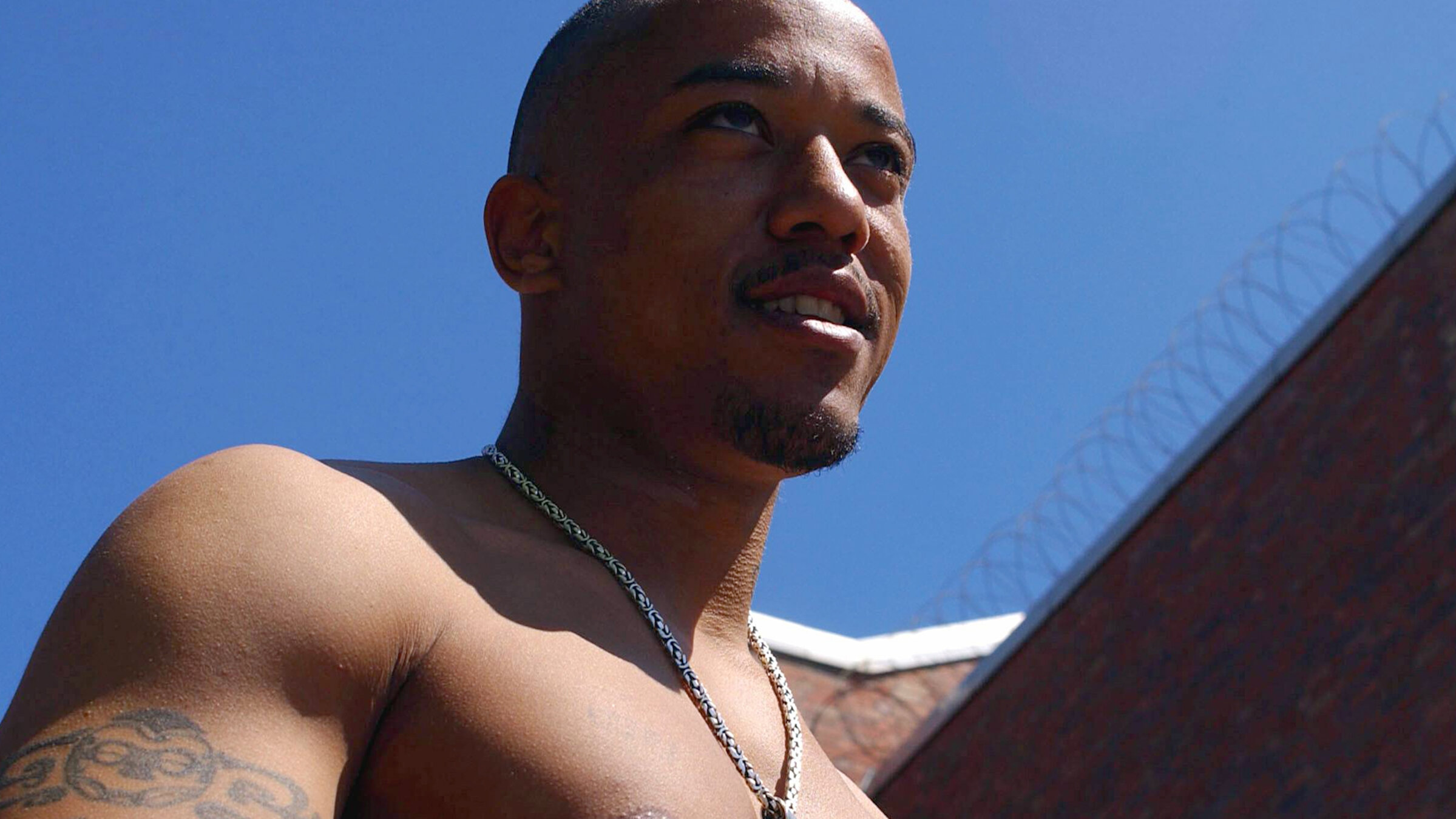 "Die Honigfalle – Eine wahre Geschichte über Liebe, Lügen und das FBI": Nahaufnahme eines jungen Mannes mit nacktem Oberkörper. Im Hintergrund Gefängnismauern mit Stacheldraht.