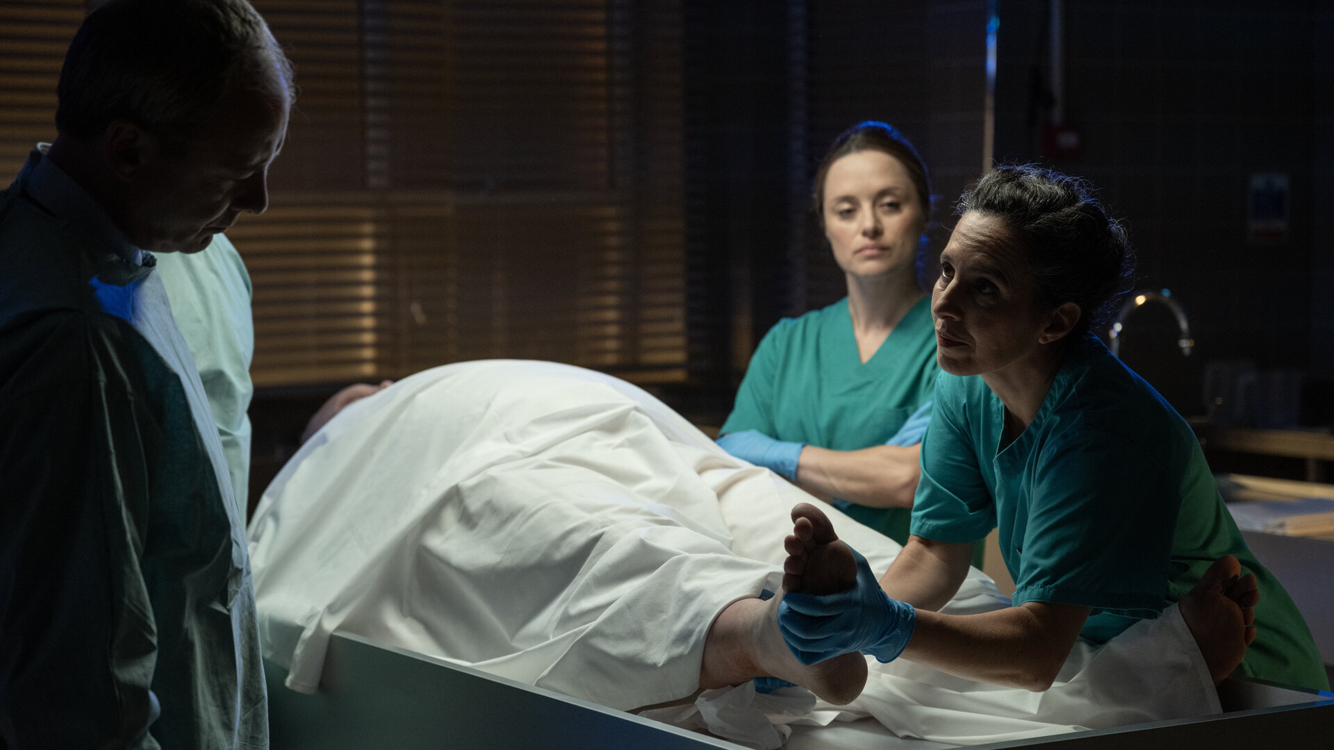 "Detective Grace - Deine Liebe ist der Tod": Roy Grace (John Simm), Cleo (Zoë Tapper) und Nadiuska De Sancha (Carolina Valdés) stehen um einen Leichentisch in der Rechtsmedizin und betrachten den Körper des toten Shelby Stoner (Daniel Fearn), der größtenteils unter einem weißen Laken verborgen ist. Nadiuska hält Shelbys rechtes Bein hoch und zeigt es Roy. Alle tragen grüne Schutzkleidung und blaue Latex-Handschuhe.