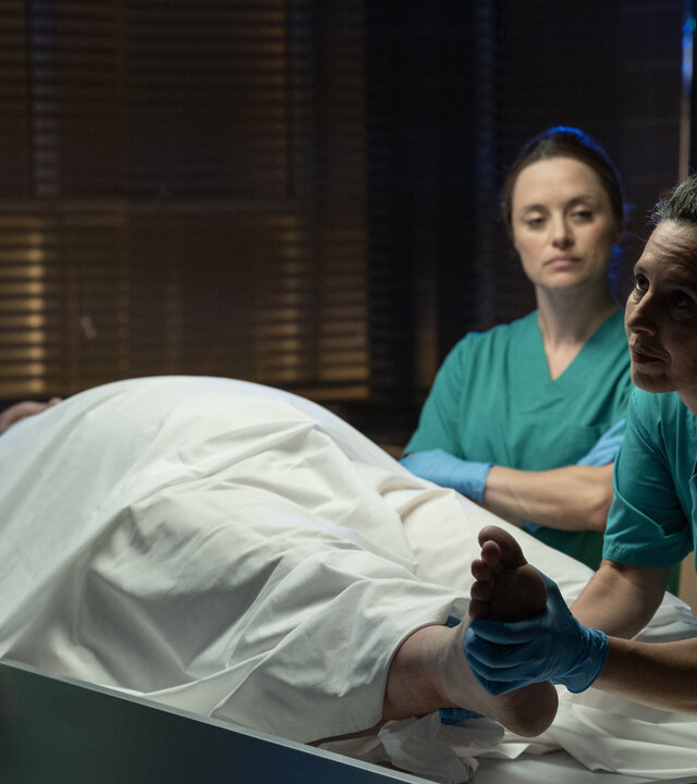 "Detective Grace - Deine Liebe ist der Tod": Roy Grace (John Simm), Cleo (Zoë Tapper) und Nadiuska De Sancha (Carolina Valdés) stehen um einen Leichentisch in der Rechtsmedizin und betrachten den Körper des toten Shelby Stoner (Daniel Fearn), der größtenteils unter einem weißen Laken verborgen ist. Nadiuska hält Shelbys rechtes Bein hoch und zeigt es Roy. Alle tragen grüne Schutzkleidung und blaue Latex-Handschuhe.