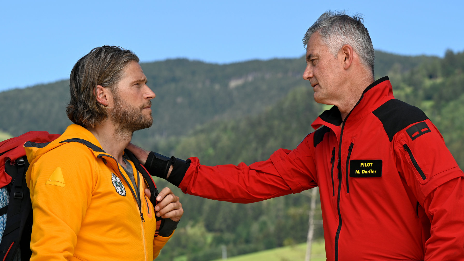 "Die Bergretter - Schamlos (1)": Auf einer Bergwiese stehen sich Bergretter Markus (Sebastian Ströbel) und Michi (Robert Lohr) gegenüber. Dieser hat seine Hand auf Markus Schulter gelegt. Beide Männer blicken sich traurig an.