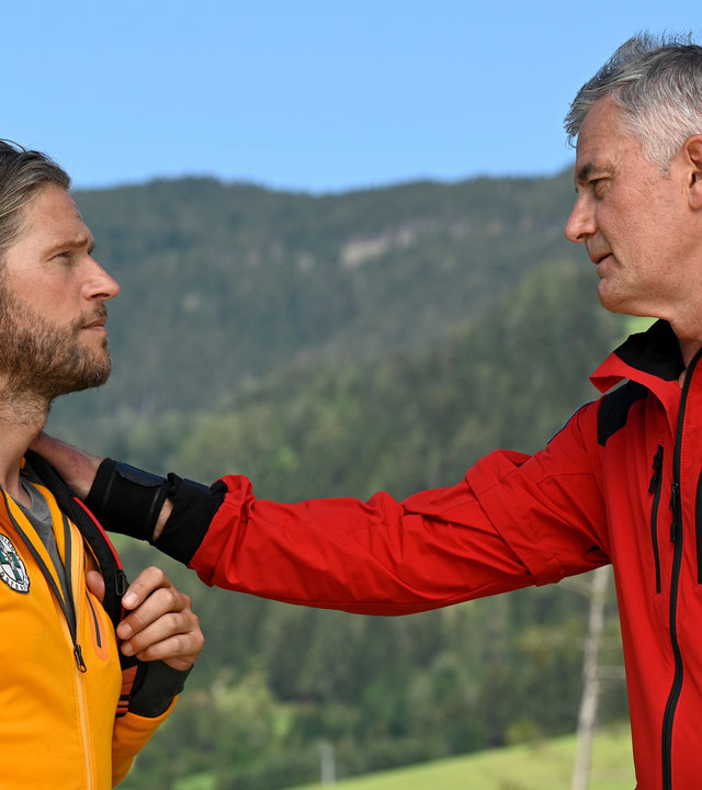 "Die Bergretter - Schamlos (1)": Auf einer Bergwiese stehen sich Bergretter Markus (Sebastian Ströbel) und Michi (Robert Lohr) gegenüber. Dieser hat seine Hand auf Markus Schulter gelegt. Beide Männer blicken sich traurig an.
