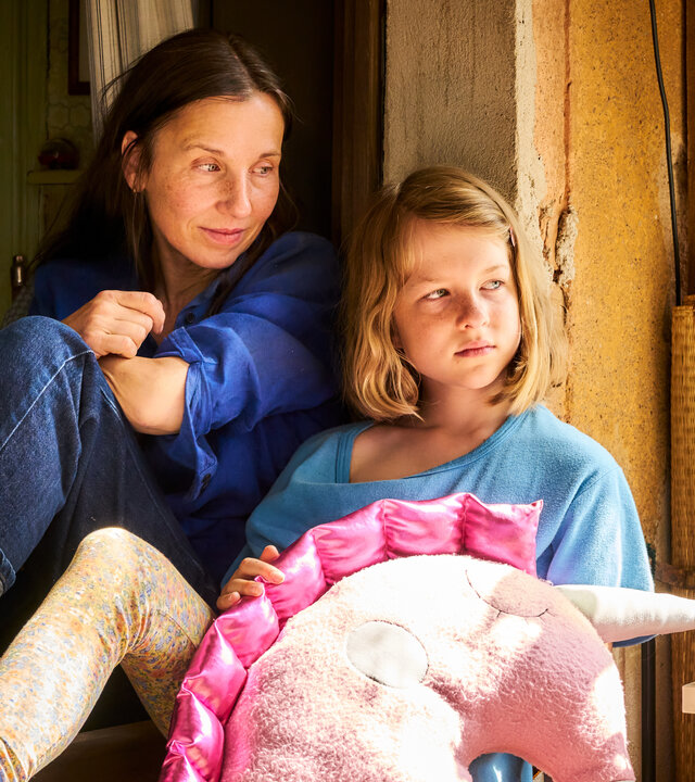 "Familie is nich": Tilda (Luise Landau) und Anne (Meret Becker) sitzen in einem alten Fensterrahmen. Putz bröckelt von der gelb gestrichenen Wand. Tilda hat ein großes Kuschelkissen in Form eines Einhornkopfs auf dem Schoß und schaut zur Seite. Anne schaut liebevoll zu Tilda.
