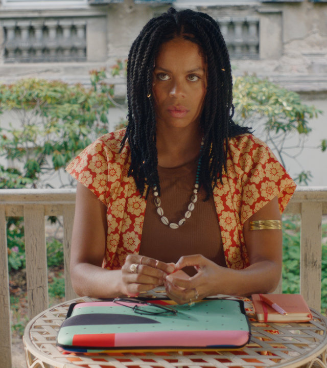"Clashing Differences – #WIR GEHEN MIT DEM TREND": Flora (Jane Chirwa) sitzt an einem kleinen runden Tisch in einem Holzpavillon und schaut in die Ferne. Auf dem Tisch liegen ein Laptop und ein Notizbuch mit einem Stift.