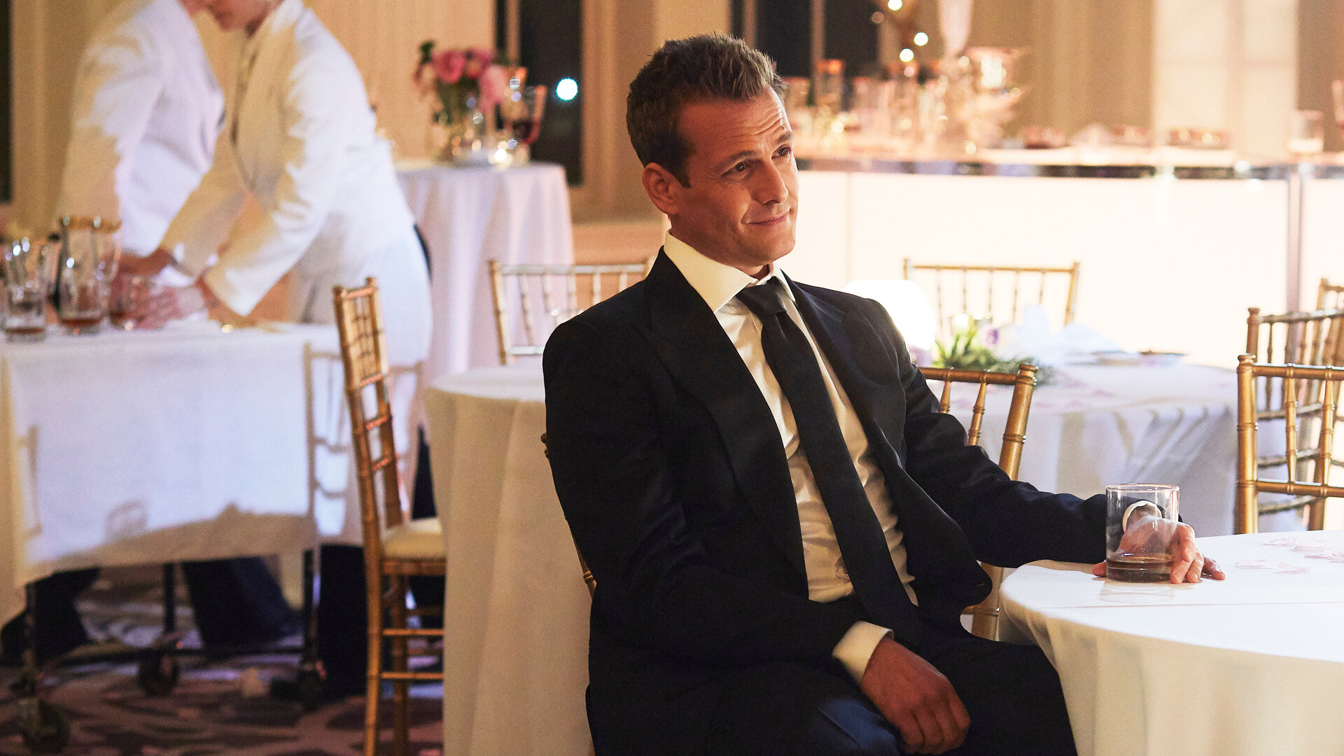 "Suits - Die rechte Hand": Harvey (Gabriel Macht) lächelt und sitzt an einem festlich eingedeckten Tisch. In seiner linken Hand, die auf dem Tisch liegt, hält er ein Whiskeyglas. Im Hintergrund sind zwei arbeitende Servicekräfte zu sehen.