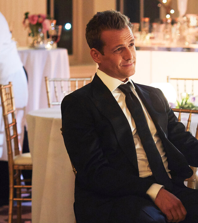 "Suits - Die rechte Hand": Harvey (Gabriel Macht) lächelt und sitzt an einem festlich eingedeckten Tisch. In seiner linken Hand, die auf dem Tisch liegt, hält er ein Whiskeyglas. Im Hintergrund sind zwei arbeitende Servicekräfte zu sehen.