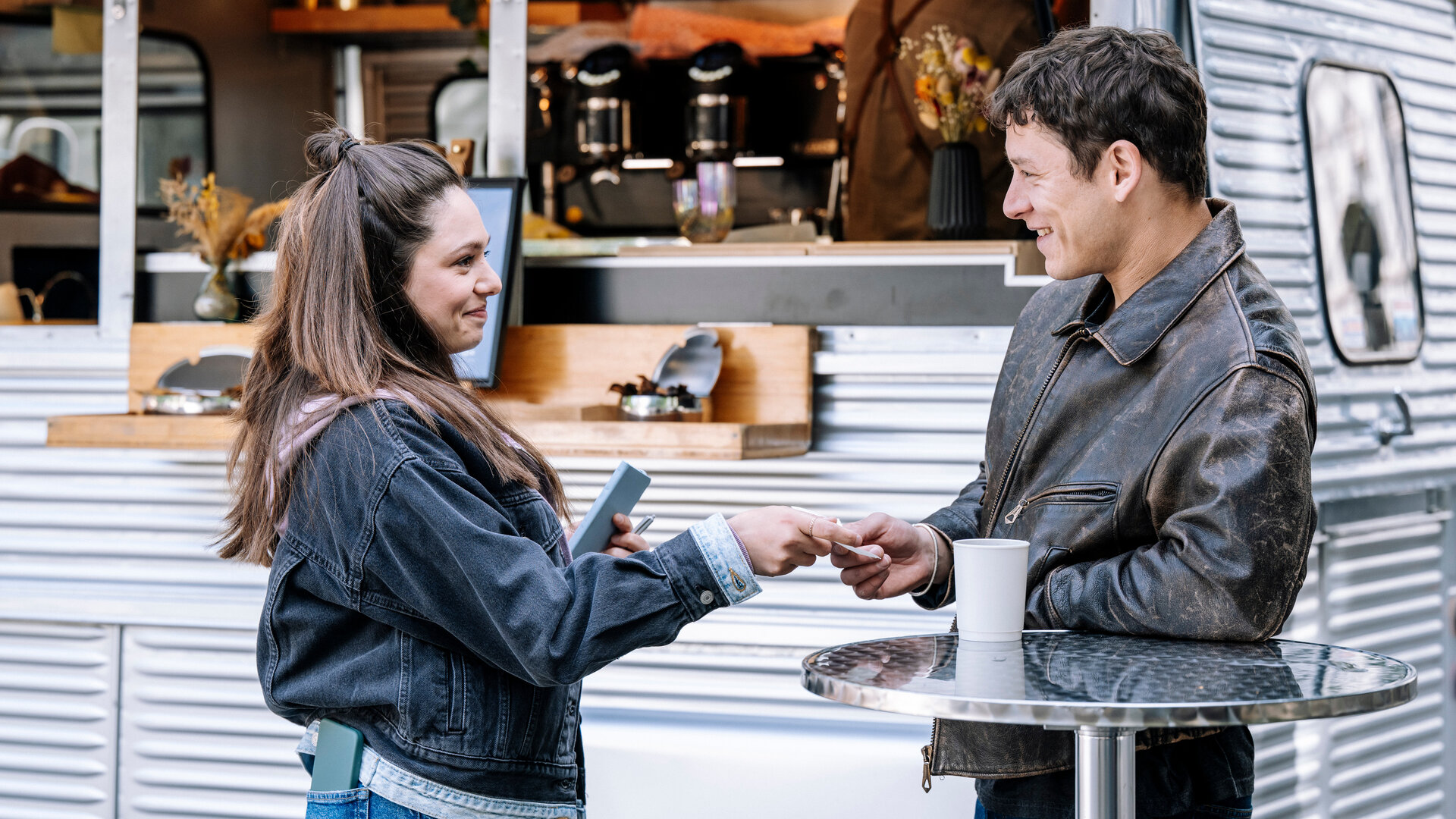 "SOKO Köln - Amiras Entscheidung": Amira Mahdi (Aylin Ravanyar) steht Max Röder (Malik Blumenthal) vor einem Food-Truck gegenüber und reicht ihm lächelnd einen Zettel. In der linken Hand hält sie ihr Handy. Max hat sich mit dem Arm auf einem silbernen Stehtisch abgestützt und erwidert ihr Lächeln. Neben ihm auf dem Tisch steht ein weißer Becher.