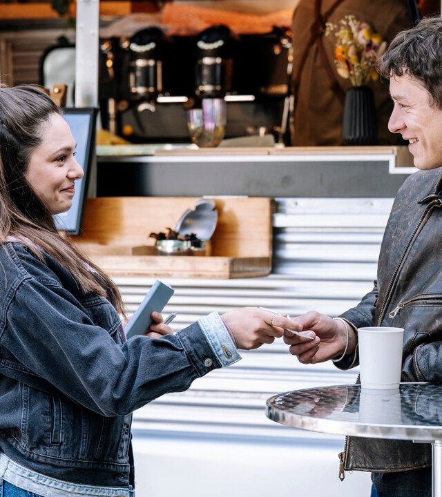 "SOKO Köln - Amiras Entscheidung": Amira Mahdi (Aylin Ravanyar) steht Max Röder (Malik Blumenthal) vor einem Food-Truck gegenüber und reicht ihm lächelnd einen Zettel. In der linken Hand hält sie ihr Handy. Max hat sich mit dem Arm auf einem silbernen Stehtisch abgestützt und erwidert ihr Lächeln. Neben ihm auf dem Tisch steht ein weißer Becher.