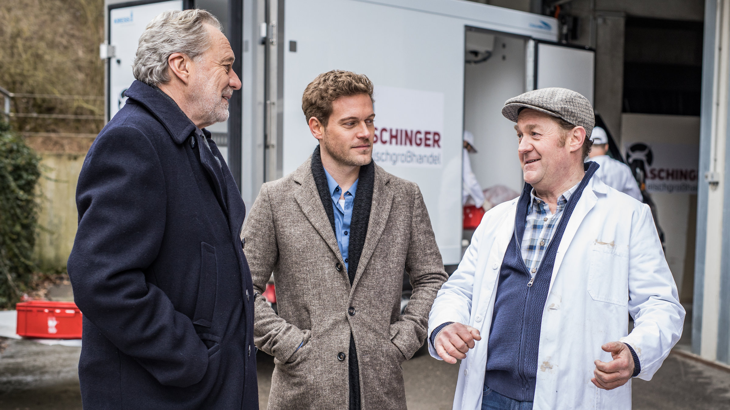 "SOKO München - Tödliche Vergangenheit": Die zwei Kommissare Arthur Bauer (Gerd Silberbauer) und Dominik Morgenstern (Joscha Kiefer) stehen mit einem der Giesinger Eiszapfen, Josef Aschinger (Jürgen Tonkel), vor seiner Metzgerei. Alle drei haben ein Lächeln im Gesicht. Die Kommissare haben die Hände in den Jackentaschen, während Josef Aschinger gestikuliert. Im Hintergrund ist ein Lieferwagen der Metzgerei zu sehen.