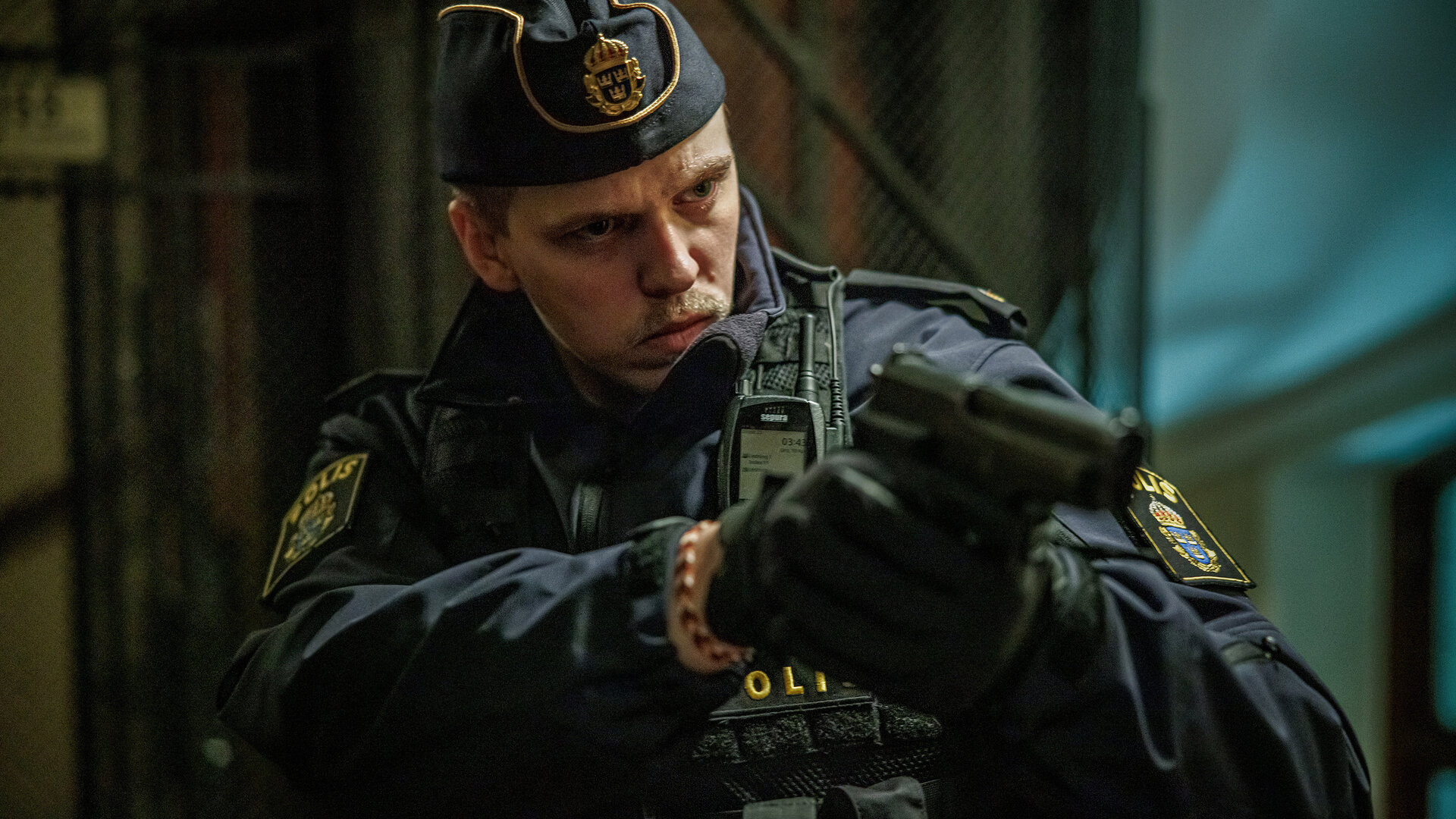 "Kommissar Beck - Vilhelm": Vilhelm Beck (Valter Skarsgård) trägt eine Polizeiuniform mitsamt Schiffchenmütze. An seiner Jacke ist eine Bodycam befestigt. Er befindet sich in einem Raum und hält mit beiden Händen eine Pistole im Anschlag.