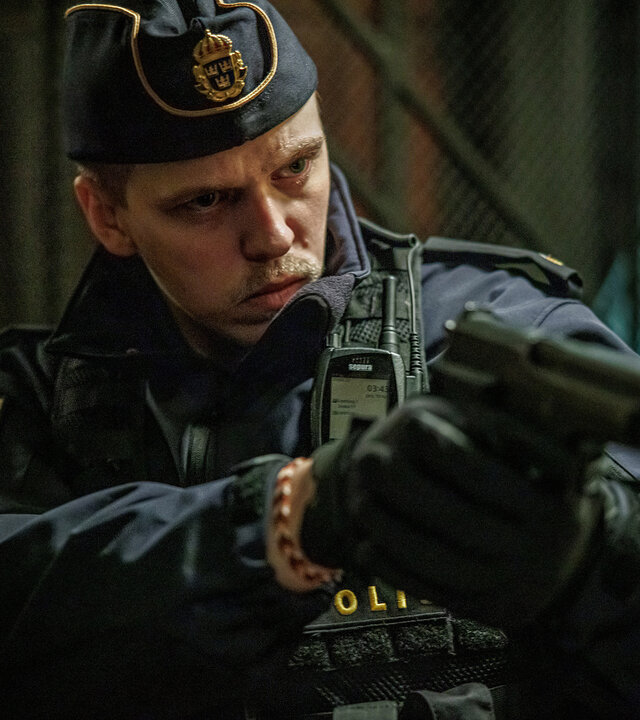 "Kommissar Beck - Vilhelm": Vilhelm Beck (Valter Skarsgård) trägt eine Polizeiuniform mitsamt Schiffchenmütze. An seiner Jacke ist eine Bodycam befestigt. Er befindet sich in einem Raum und hält mit beiden Händen eine Pistole im Anschlag.