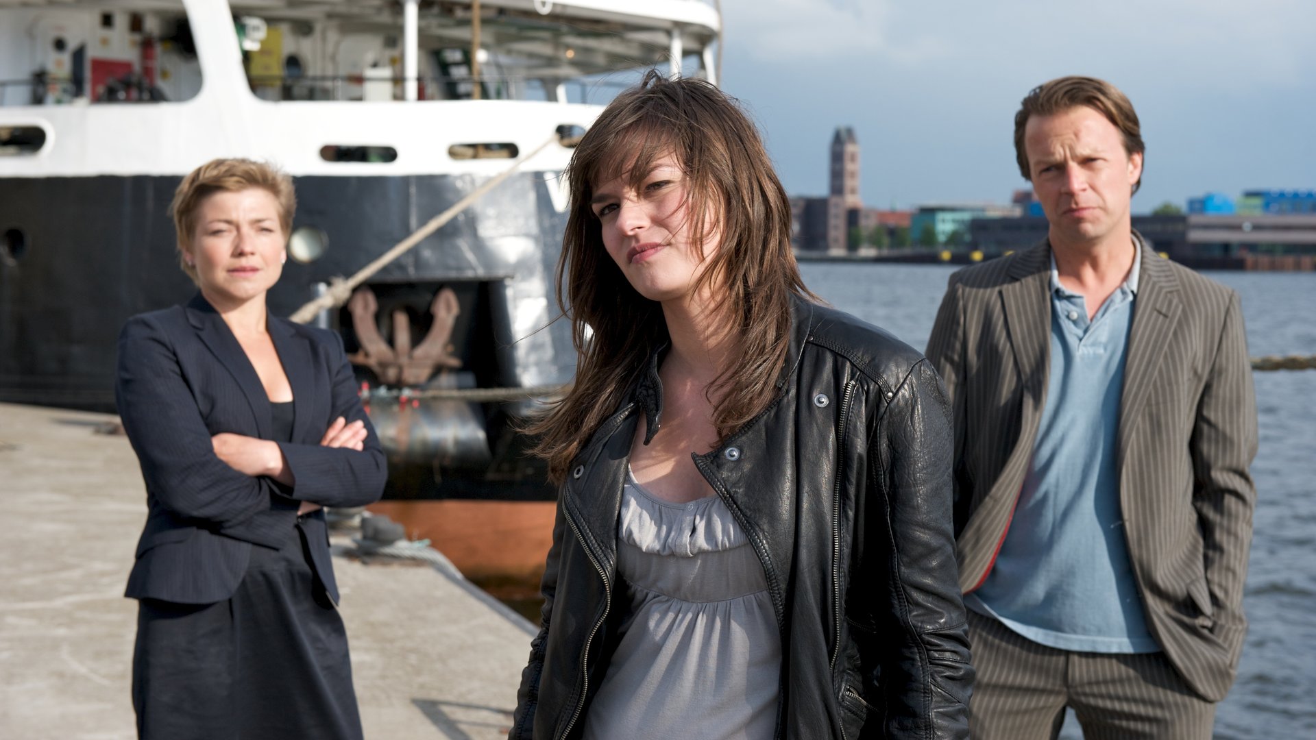 "SOKO Wismar - Der blonde Hans": Cindy Lundt (Diane Willems) steht mit dem Rücken zu den Ermittlern Katrin Börensen (Claudia Schmutzler) und Sven Herzog (Michael Härle) im Hafen von Wismar. Im Hintergrund ist ein Schiff vertäut, und die Stadt sowie der Hafen sind zu erkennen.