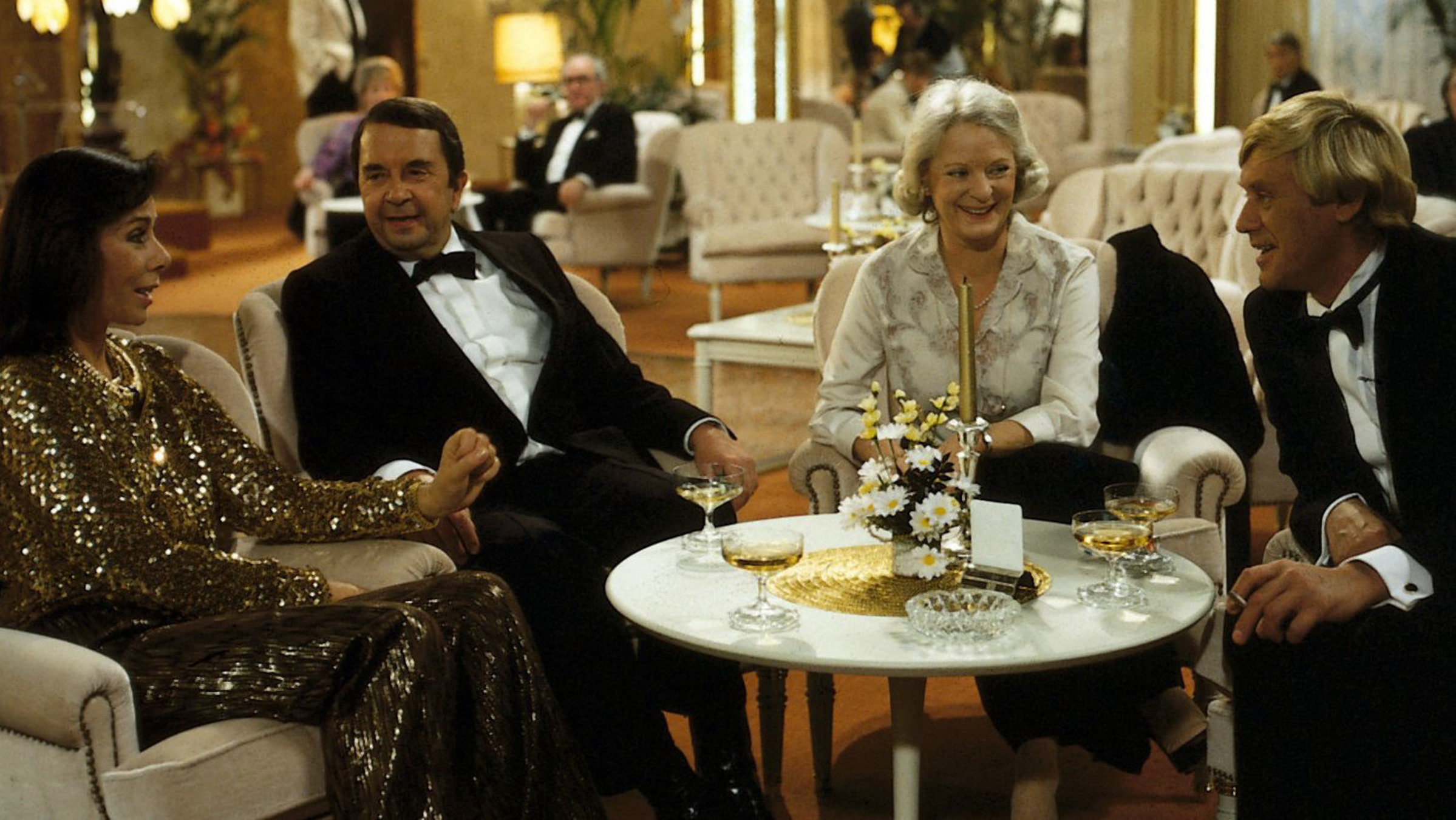 "Das Traumschiff": Das Ehepaar Borchert (Elisabeth Wiedemann, Wolfgang Kieling) sitzen mit dem Ehepaar Neumann (Monika Peitsch, Horst Frank) in einem Schiffssalon und trinken Wein. Sie unterhalten sich fröhlich.