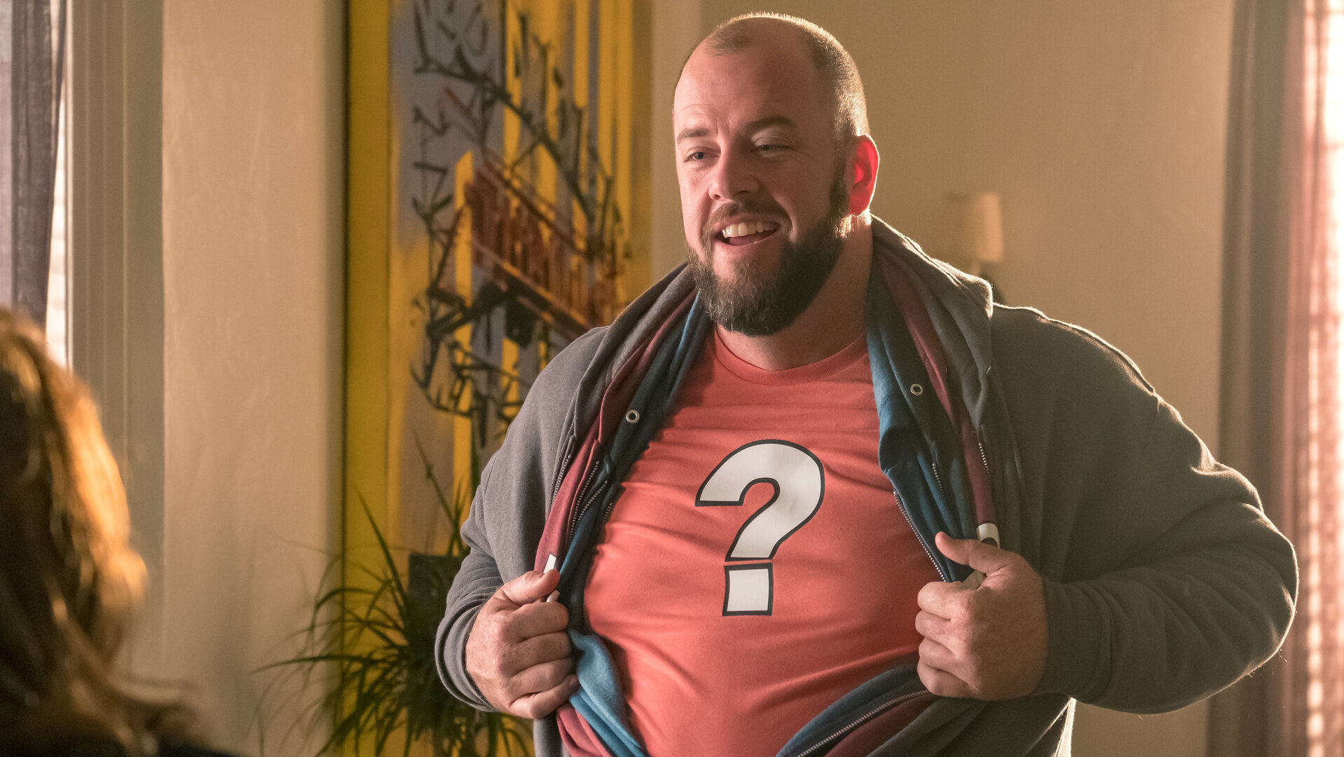 "This Is Us - Enttäuschungen": Toby Damon (Chris Sullivan) steht in einem Raum. Er trägt mehrere farbige Sweatshirt‑Jacken übereinander und zieht sie auseinander. Dabei zeigt er ein rotes T‑Shirt mit einem großen weißen Fragezeichen, das er unter all den Jacken trägt. Er blickt lächelnd nach vorn.