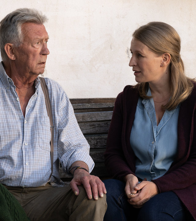 "Die Bergretter: Klang der Erinnerung (2)": Franz (Heinz Marecek) sitzt auf einer Holzbank und blickt entsetzt zu Emilie (Stefanie von Poser), die neben ihm sitzt.