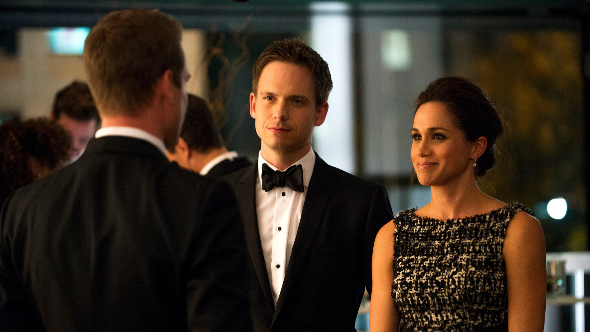 "Suits - Krieg": Mike (Patrick J. Adams) und Rachel (Meghan Markle) stehen nebeneinander und blicken lächelnd zu Harvey (Gabriel Macht), der ihnen gegenübersteht. Alle drei tragen festliche Abendgarderobe.