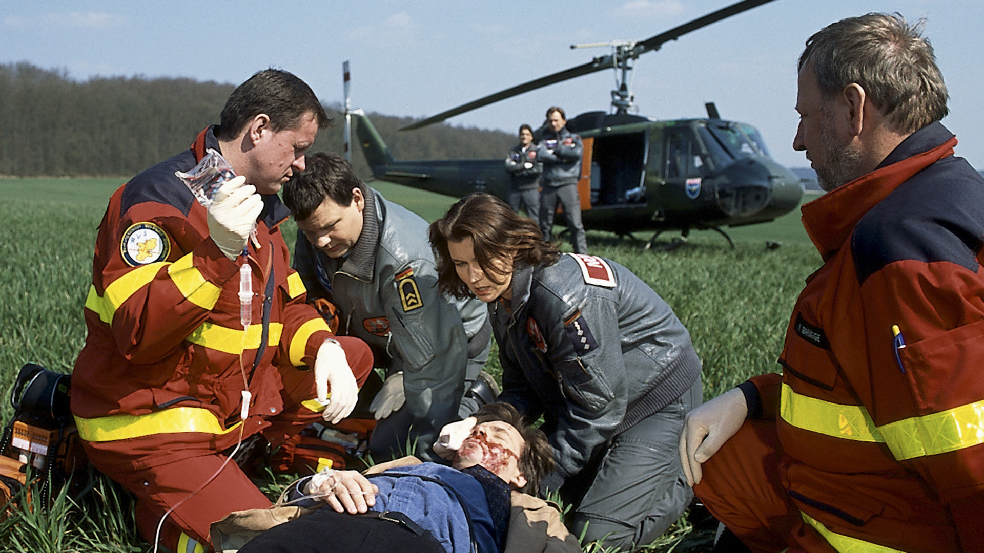 "Die Rettungsflieger - Wollckes Wagnis": Slawomir (Tomek Nowicki) liegt schwer verletzt auf einer Wiese. Ilona (Julia Heinemann) und Thomas (Ulrich Bähnk) knien zusammen mit zwei Sanitätern neben ihm und versorgen ihn. Im Hintergrund sieht man zwei weitere Rettungsflieger und den Hubschrauber.