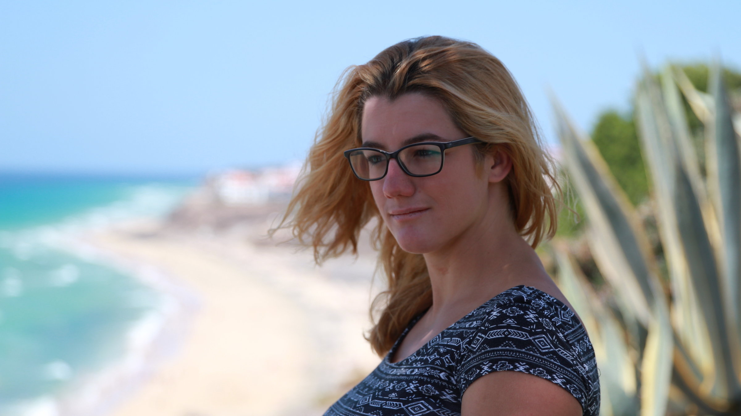"37°: Ich lebe positiv - Corinne und ihr Schicksal HIV":  Corinne steht an einem gelben Sandstrand. Das Meer ist hellblau und hinter ihr ist eine große Agave zu sehen.