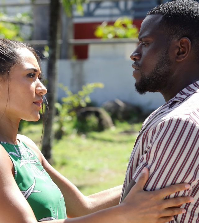 "Death in Paradise- Geheimnisvolle Tiefen - Teil 1": Auf einer sonnigen Straße. Florence Cassell (Joséphine Jobert) und Patrice Campbell (Leemore Marrett Jr.) schauen sich verliebt in die Augen.