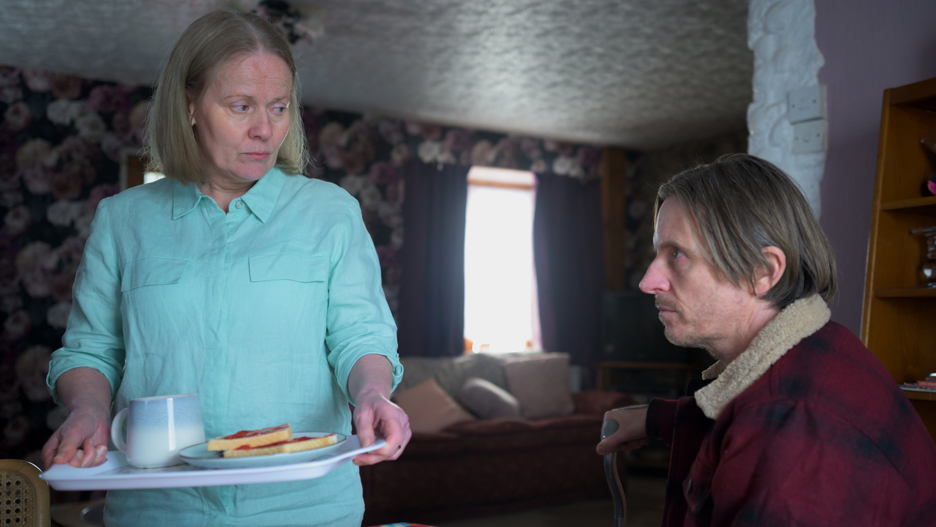 "Die Pembrokeshire Morde (2)": Adrian (Oliver Ryan) sitzt an einem Tisch und sieht zu seiner Mutter Pat (Caroline Berry), die neben ihm steht. Sie trägt ein Tablett mit einer Tasse und zwei Marmeladentoasts in den Händen und erwidert seinen Blick.