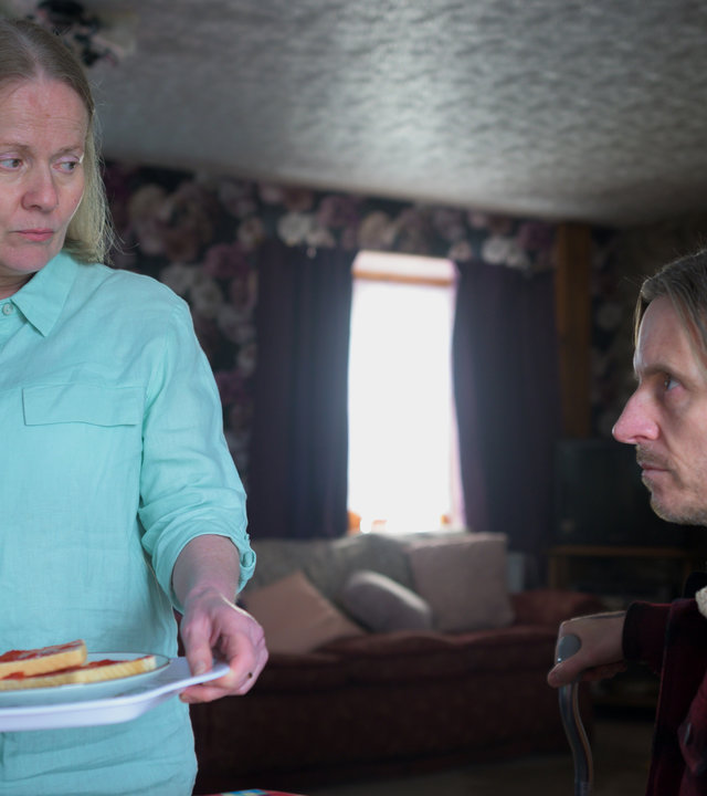 "Die Pembrokeshire Morde (2)": Adrian (Oliver Ryan) sitzt an einem Tisch und sieht zu seiner Mutter Pat (Caroline Berry), die neben ihm steht. Sie trägt ein Tablett mit einer Tasse und zwei Marmeladentoasts in den Händen und erwidert seinen Blick.