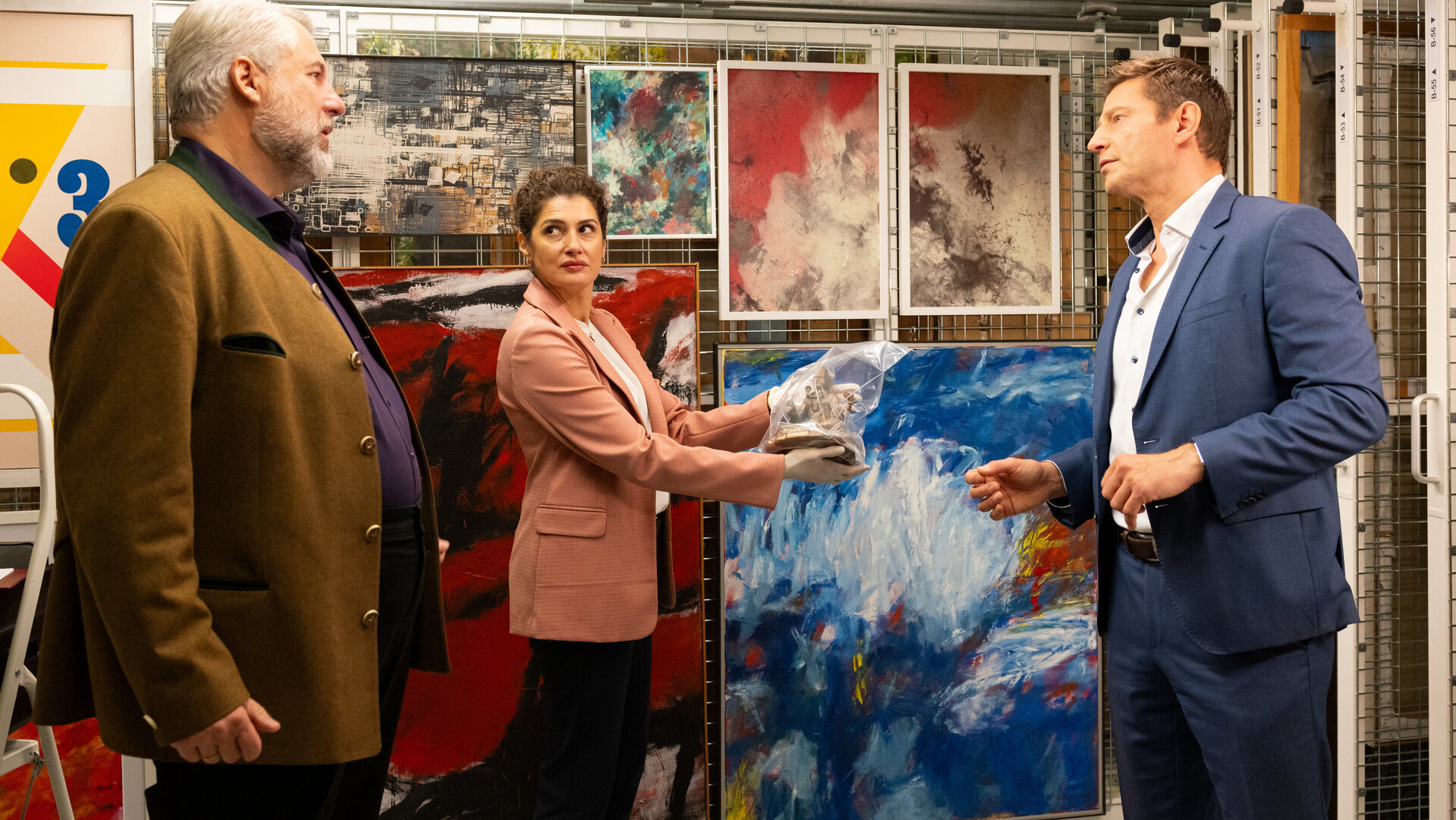 "Die Rosenheim-Cops - Ein Schwarm für Hansen": Kommissar Stadler (Dieter Fischer), Rechtsmedizinerin Dr. Dimos (Anastasia Papadopoulou) und Kommissar Hansen (Igor Jeftić) stehen um das Opfer (Komparsin) herum, das auf dem Boden liegt. Im Hintergrund hängen diverse moderne Gemälde.