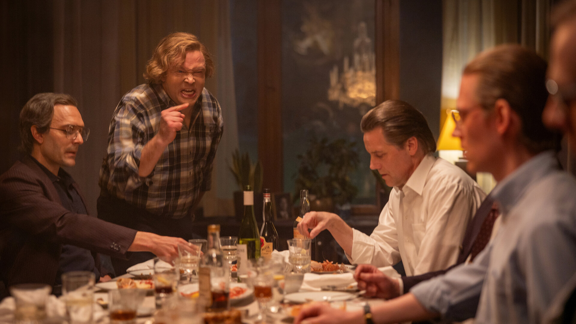 "Stenbeck - Noch ein Bissen - Macht, Medien und Monopole": Jan Stenbeck (Jakob Oftebro) steht am Kopfende eines gedeckten Tisches und redet wutentbrannt auf einen der vor ihm sitzenden Männer ein.