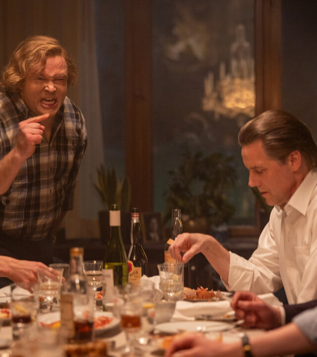 "Stenbeck - Noch ein Bissen - Macht, Medien und Monopole": Jan Stenbeck (Jakob Oftebro) steht am Kopfende eines gedeckten Tisches und redet wutentbrannt auf einen der vor ihm sitzenden Männer ein.