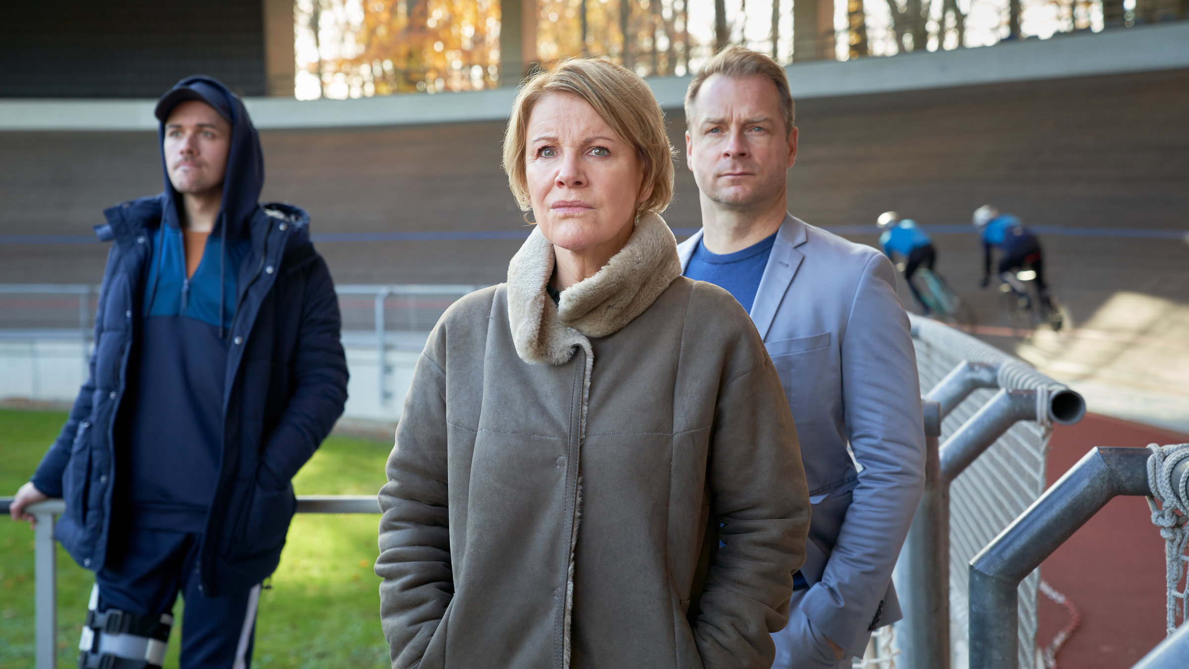 "Marie Brand und der Tote im Trikot": Marie Brand (Mariele Millowitsch) und Jürgen Simmel (Hinnerk Schönemann) stehen mit Tharde Rickling (Fred Costea) im Bahnrad-Station. Tharde Rickling hält sich mit dem rechten Arm an einem Geländer fest. Marie Brand und Jürgen Simmel haben jeweils ihre Hände in den Jackentaschen. Im Hintergrund sieht man Radfahrer auf der Bahn trainieren.