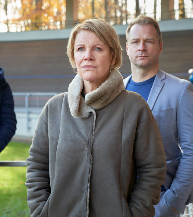 "Marie Brand und der Tote im Trikot": Marie Brand (Mariele Millowitsch) und Jürgen Simmel (Hinnerk Schönemann) stehen mit Tharde Rickling (Fred Costea) im Bahnrad-Station. Tharde Rickling hält sich mit dem rechten Arm an einem Geländer fest. Marie Brand und Jürgen Simmel haben jeweils ihre Hände in den Jackentaschen. Im Hintergrund sieht man Radfahrer auf der Bahn trainieren.