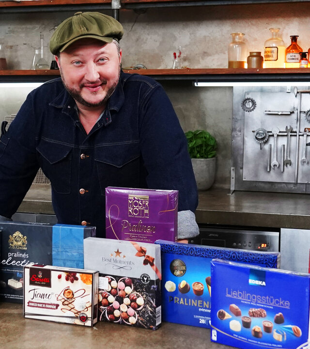 "besseresser: Falsche Feinkost-Versprechen - Sebastian Lege packt aus": Sebastian Lege steht hinter verschiedenen Pralinen-Verpackungen.