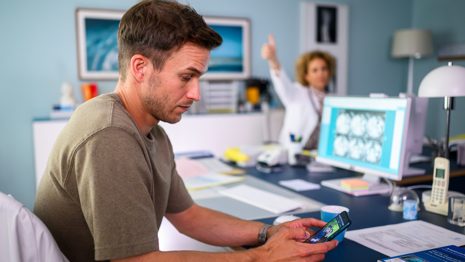 "Bettys Diagnose - Herzenssache": Dr. Paul Eckart (Florian Frowein) sitzt am Schreibtisch und schaut auf sein Smartphone. Im Hintergrund ist Dr. Helena von Arnstett (Claudia Hiersche) im weißen Arztkittel zu sehen, die den Arm in seine Richtung hochhält und mit dem Daumen nach oben zeigt.