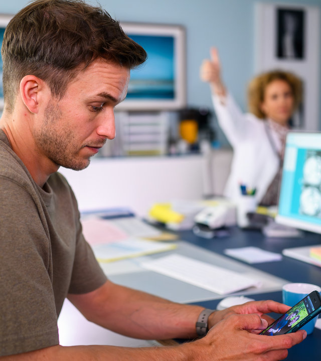 "Bettys Diagnose - Herzenssache": Dr. Paul Eckart (Florian Frowein) sitzt am Schreibtisch und schaut auf sein Smartphone. Im Hintergrund ist Dr. Helena von Arnstett (Claudia Hiersche) im weißen Arztkittel zu sehen, die den Arm in seine Richtung hochhält und mit dem Daumen nach oben zeigt.