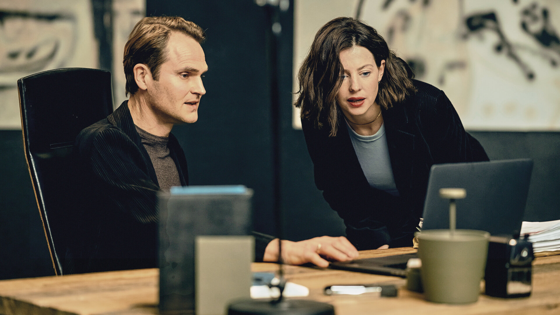 "Jugend – es ist kompliziert: Die Neue": Florian Graf (Fabian Hinrichs) sitzt in einem schwarzen Sakko an einem Bürotisch, Cathrin (Sarah Gailer) steht daneben, beide schauen auf ein Laptop.
