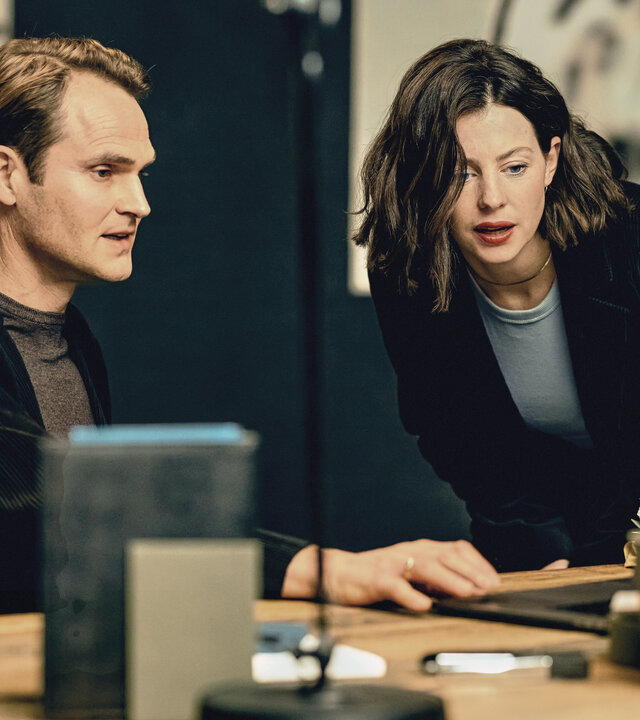 "Jugend – es ist kompliziert: Die Neue": Florian Graf (Fabian Hinrichs) sitzt in einem schwarzen Sakko an einem Bürotisch, Cathrin (Sarah Gailer) steht daneben, beide schauen auf ein Laptop.
