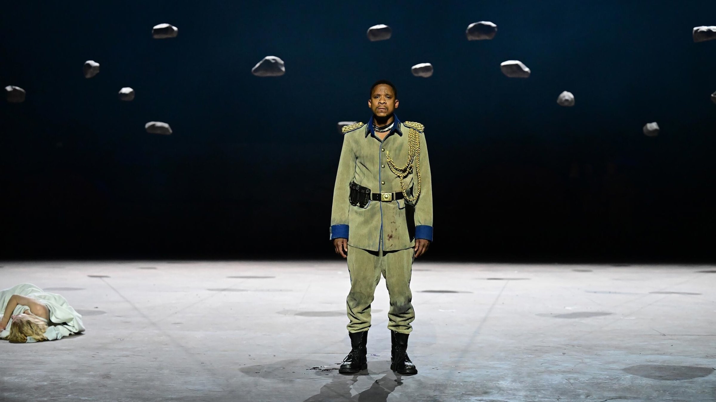 "Othello aus Südafrika – Shakespeare am Düsseldorfer Schauspielhaus": In der Mitte des Bildes steht ein Mann auf einer Bühne. Über ihm hängen an der Decke Steine an Fäden. Der Mann trägt eine grüne Soldatenuniform. Links am Bildrand liegt eine Frau auf dem Boden.