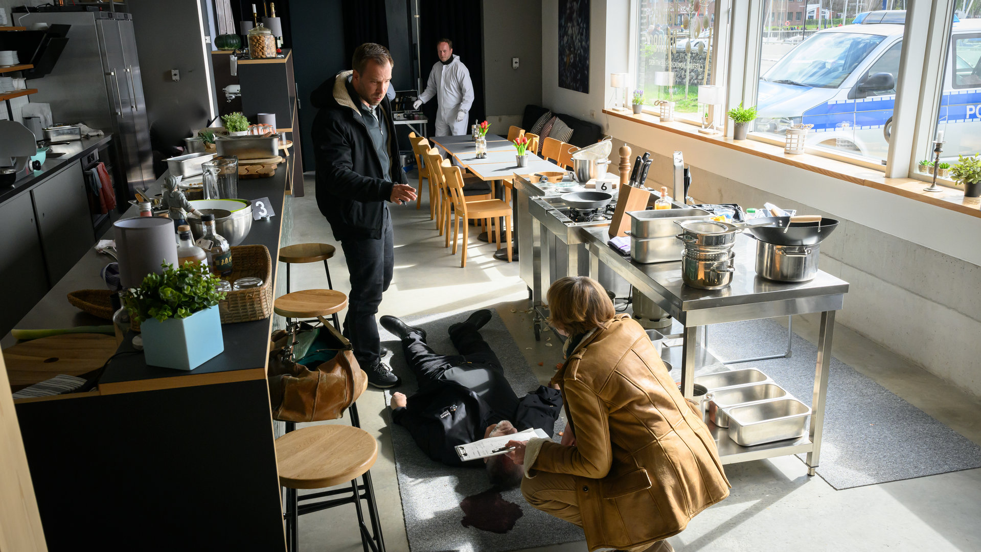 "SOKO Wismar - Haukes Dinner": Dr. Helene Sturbeck (Katharina Blaschke) kniet in der Küche vor dem toten Hauke Kahnemann (Marcus Kaloff). Lars Pöhlmann (Dominic Boeer) hat einige Fragen an sie.