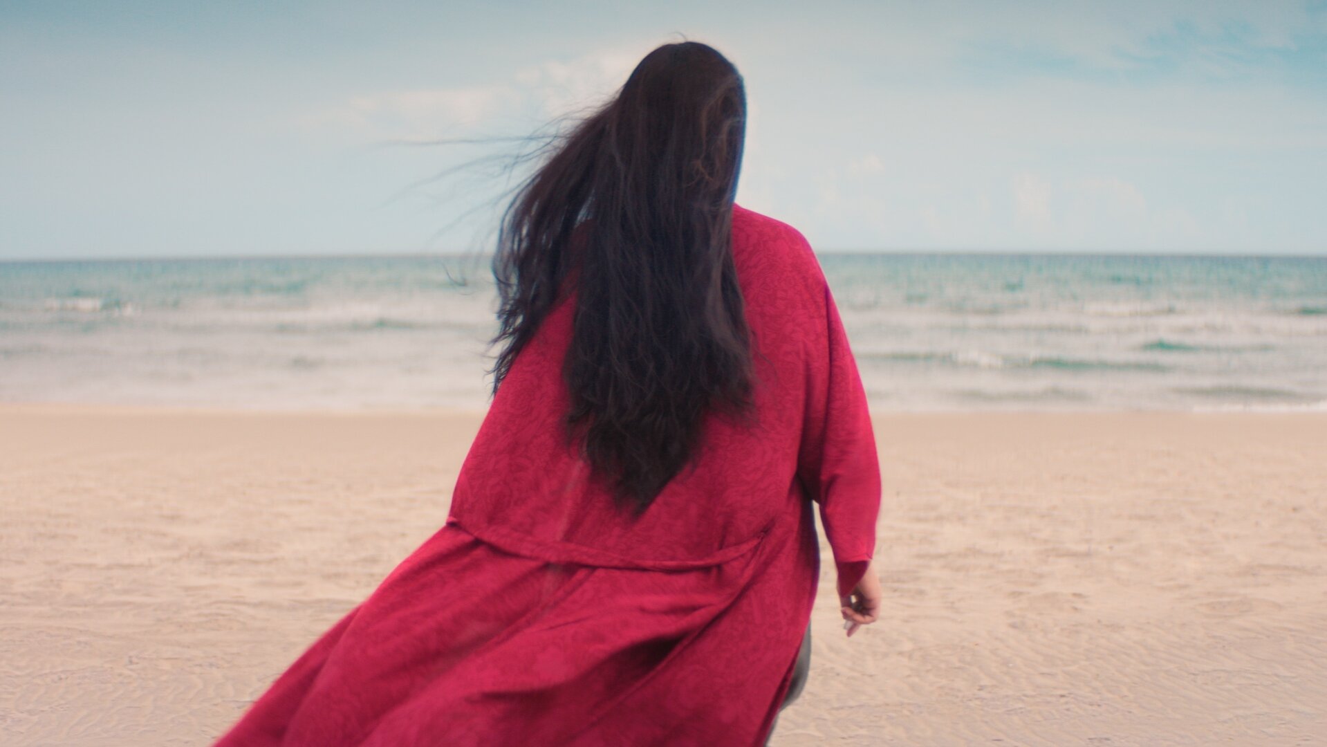 "Take the Money and Run - One Life": Ruja Ignatova (Nilam Farooq) steht in einem pinken Bademantel an einem Sandstrand und blickt auf das Meer. Sie ist nur von hinten zu sehen und ihre dunklen, offenen Haare wehen im Wind.