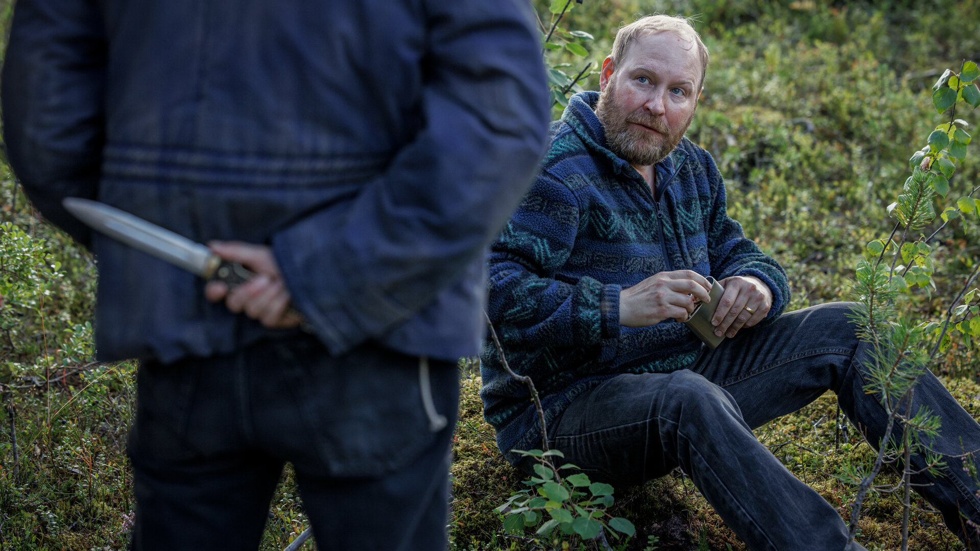 "Wolfssommer – Blutige Spuren - Der Hinterhalt": Thomas (Henrik Dorsin) sitzt im Grünen auf dem Boden. Er hält eine flache Schnapsflasche in den Händen. Ihm gegenüber steht Kat (Eliot Sumner), die nur zum Teil und von hinten zu sehen ist. Sie versteckt ein Messer hinter ihrem Rücken.